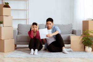 Young couple sitting on the living room floor among unpacked moving boxes, looking worried at a document they’re reading together.