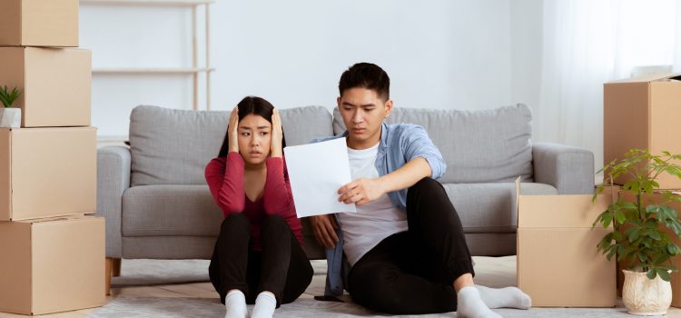 Young couple sitting on the living room floor among unpacked moving boxes, looking worried at a document they’re reading together.